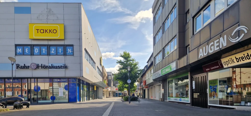 Eine Fußgängerzone in Dorf Westerholt, gesäumt von modernen Gebäuden und Geschäften, darunter Takko, MEDIZIN und AGEN, unter einem blauen Himmel mit vereinzelten Wolken. Die Straße ist bis auf einige Bänke und Skulpturen leer.
