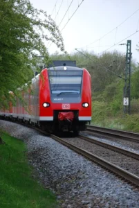 Ein roter Regionalzug der Deutschen Bahn (DB) fährt an einem bewölkten Tag in der Nähe des Bahnhofs Herten auf einem gebogenen Gleis, umgeben von grünen Bäumen und Gras.
