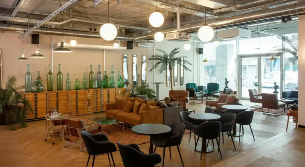 Modern eingerichteter Lounge- und Arbeitsbereich mit großen Fenstern, braunem Ledersofa, runden schwarzen Tischen, Stühlen und Sesseln. Industrieller Stil mit sichtbarer Decke, hängenden Lampen und Pflanzen, darunter Palmen und grüne Glasvasen auf einer Holzkommode.